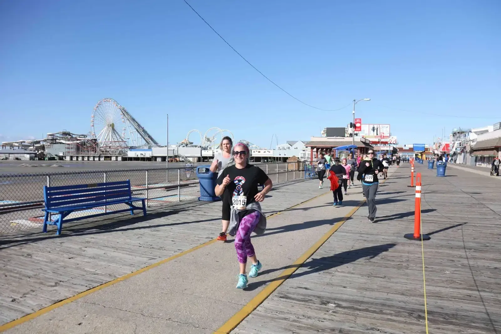 Runners participate in a race on a wooden boardwalk near the beach, with roller coasters and amusement park rides visible in the background under a clear blue sky.