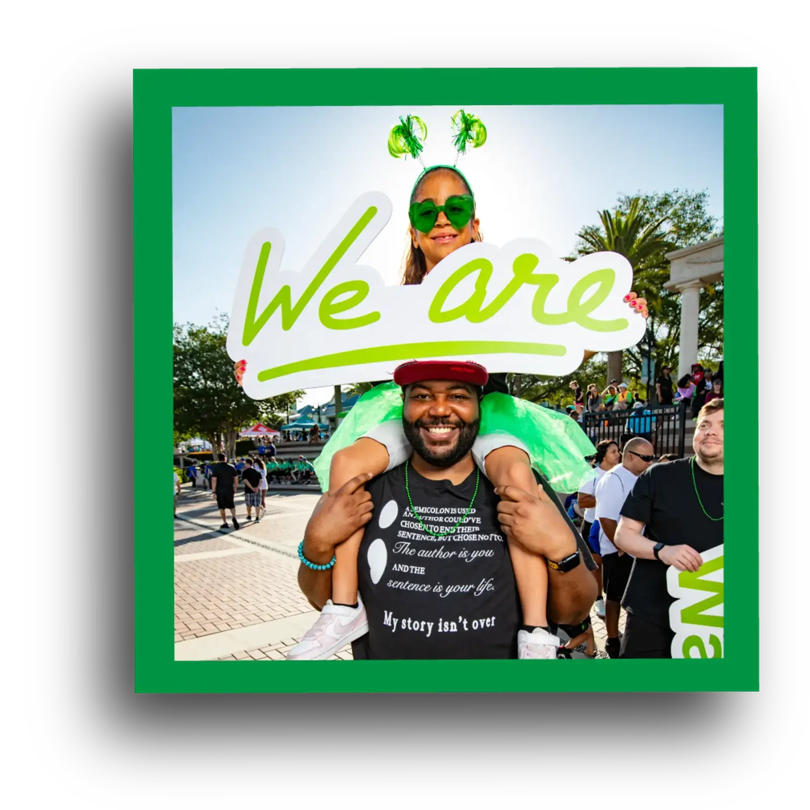 A smiling man carries a young girl on his shoulders at an outdoor event. The girl wears a green tutu and shamrock headband. Both hold a large sign reading “We are.” People and trees appear in the sunny background.