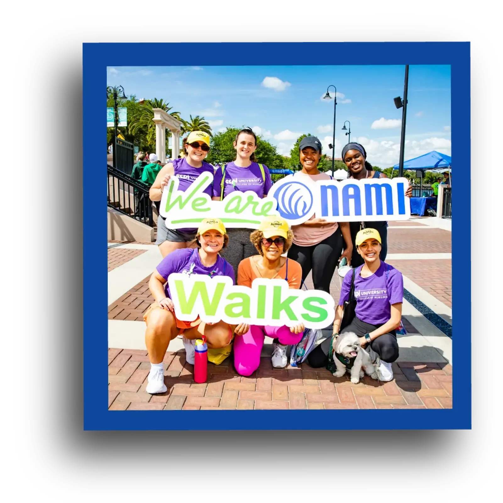 A group of seven smiling people in athletic clothes pose outdoors, holding signs that read "We are NAMI Walks," with a sunny sky and event tents in the background.
