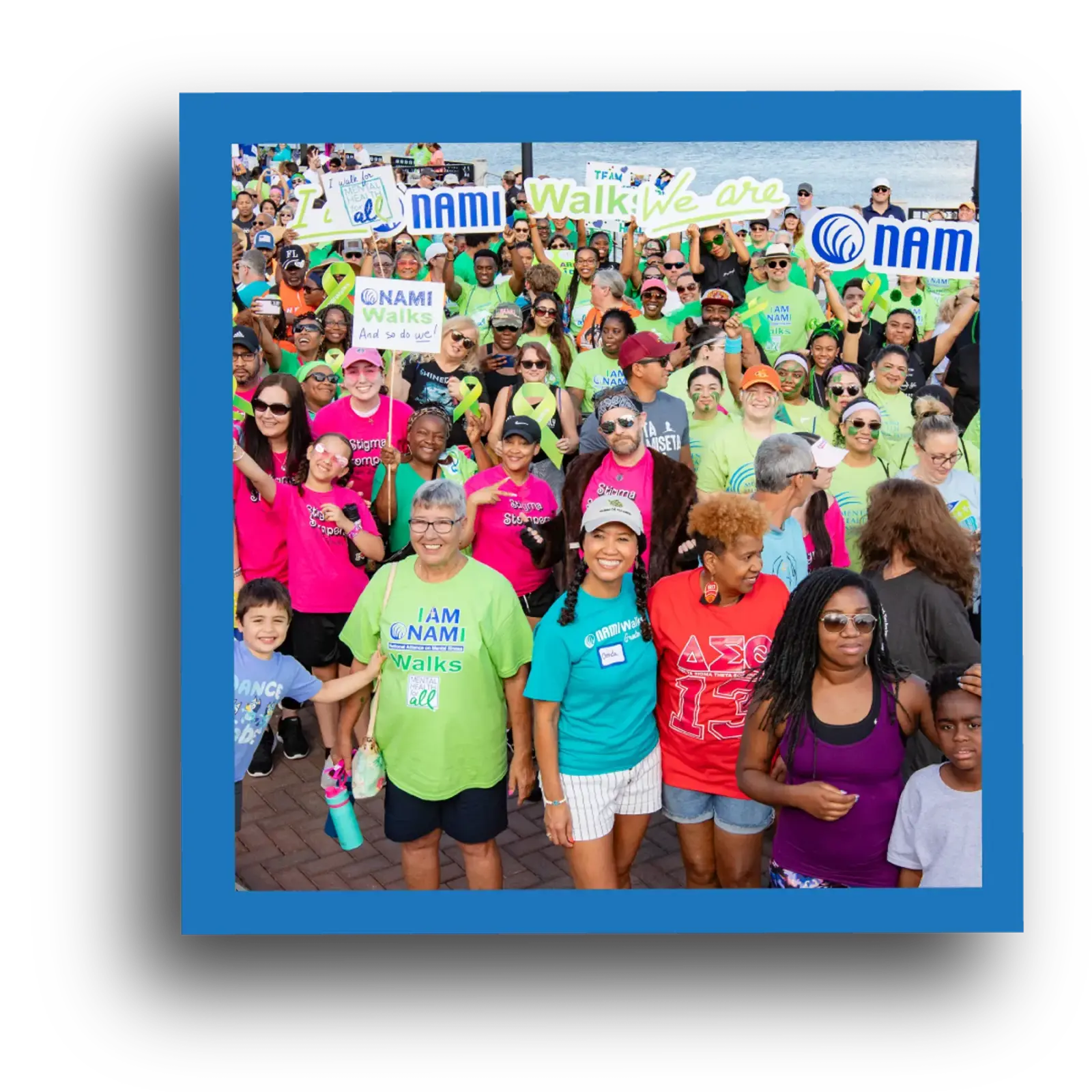 A large, diverse group of people wearing colorful shirts and sunglasses gather outdoors, smiling and holding signs for a NAMI Walk event, showing unity and support for mental health awareness.