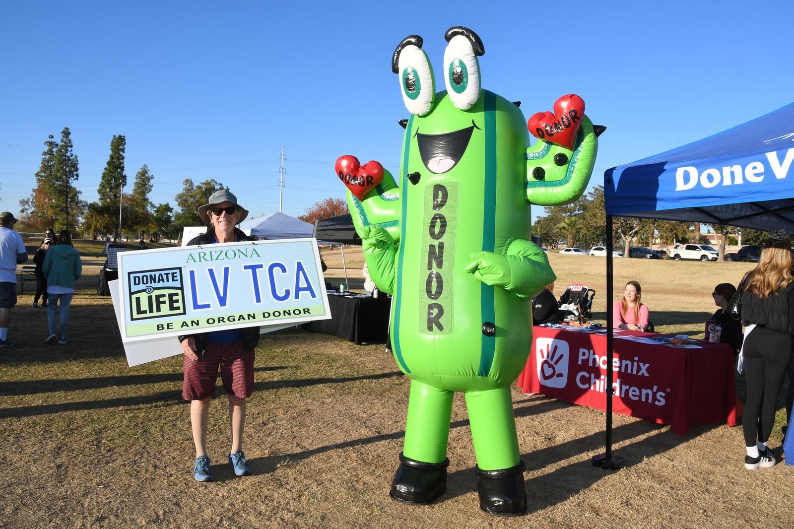A person holds an Arizona organ donor license plate sign next to a large green inflatable cactus mascot labeled "DONOR," with Phoenix Children’s booth and people in the background at an outdoor event.