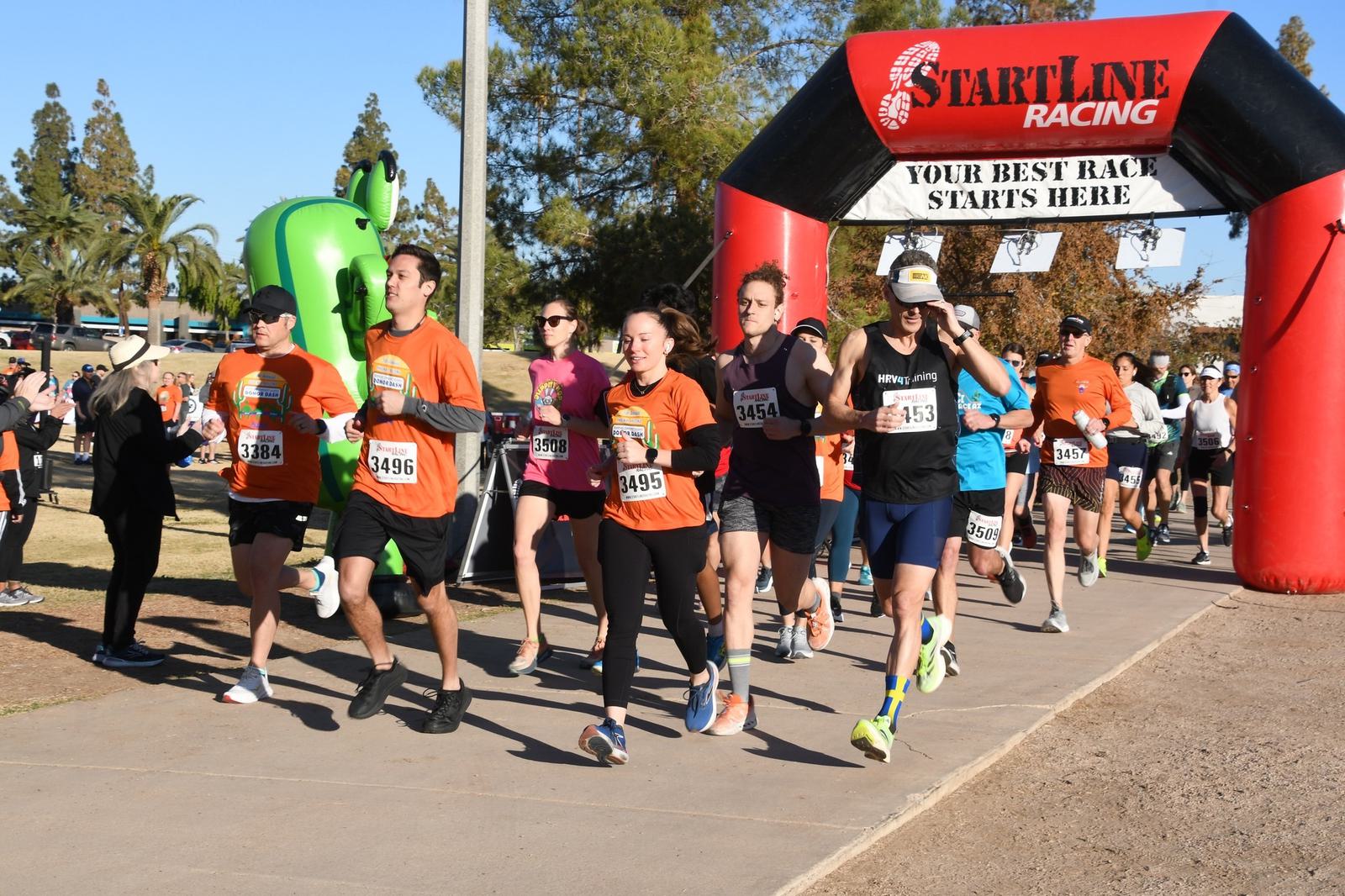 A group of runners, many wearing orange shirts, begin a race under a red and black inflatable arch that reads “START LINE RACING—YOUR BEST RACE STARTS HERE” on a sunny day. Spectators and trees are in the background.