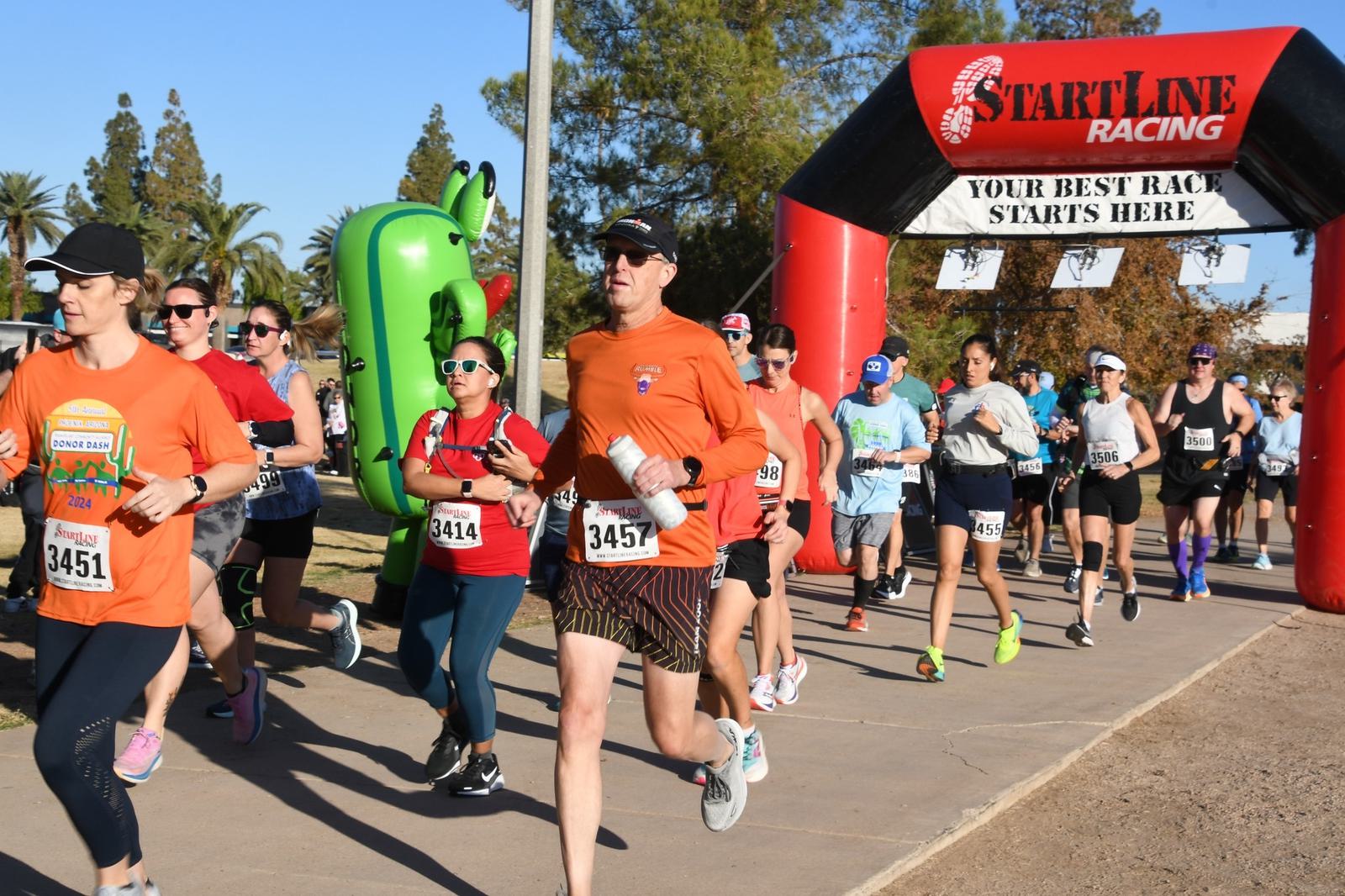 A group of runners, some in costumes, participates in a race, passing under a red and black inflatable arch that reads “StartLine Racing—Your Best Race Starts Here,” on a sunny day.