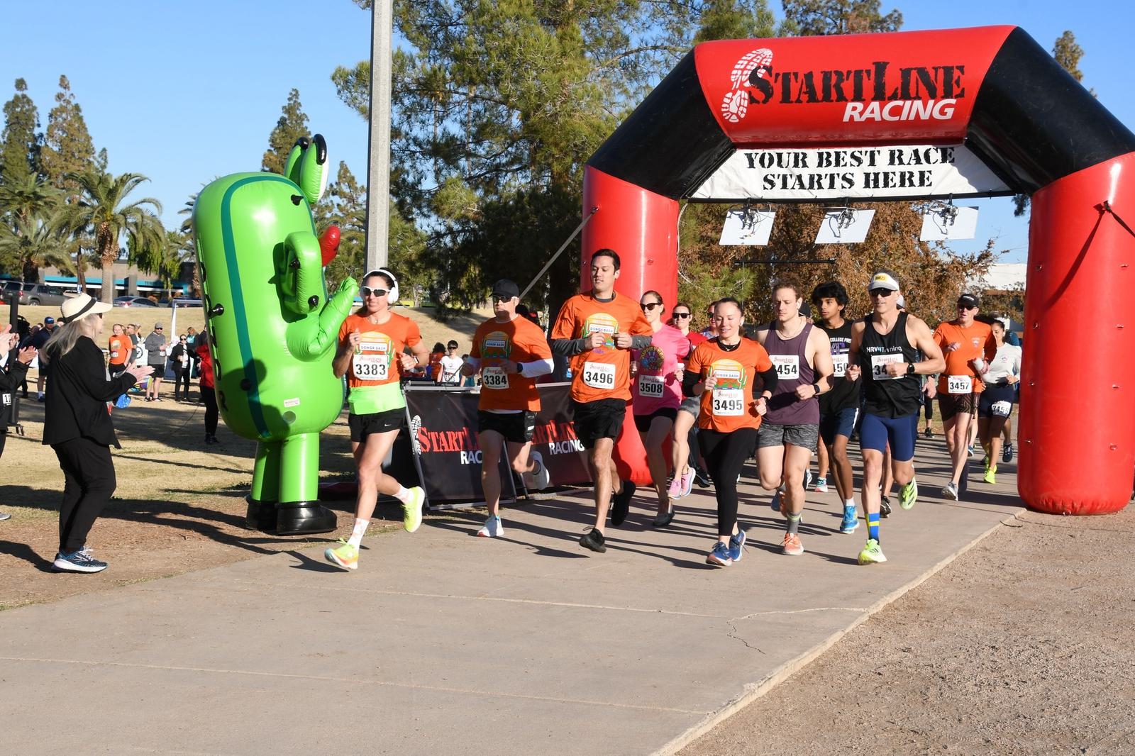 A group of runners starts a race under a red and black inflatable arch that reads “START LINE RACING.” A person in a green cactus costume is cheering beside the path. Spectators watch in the background.