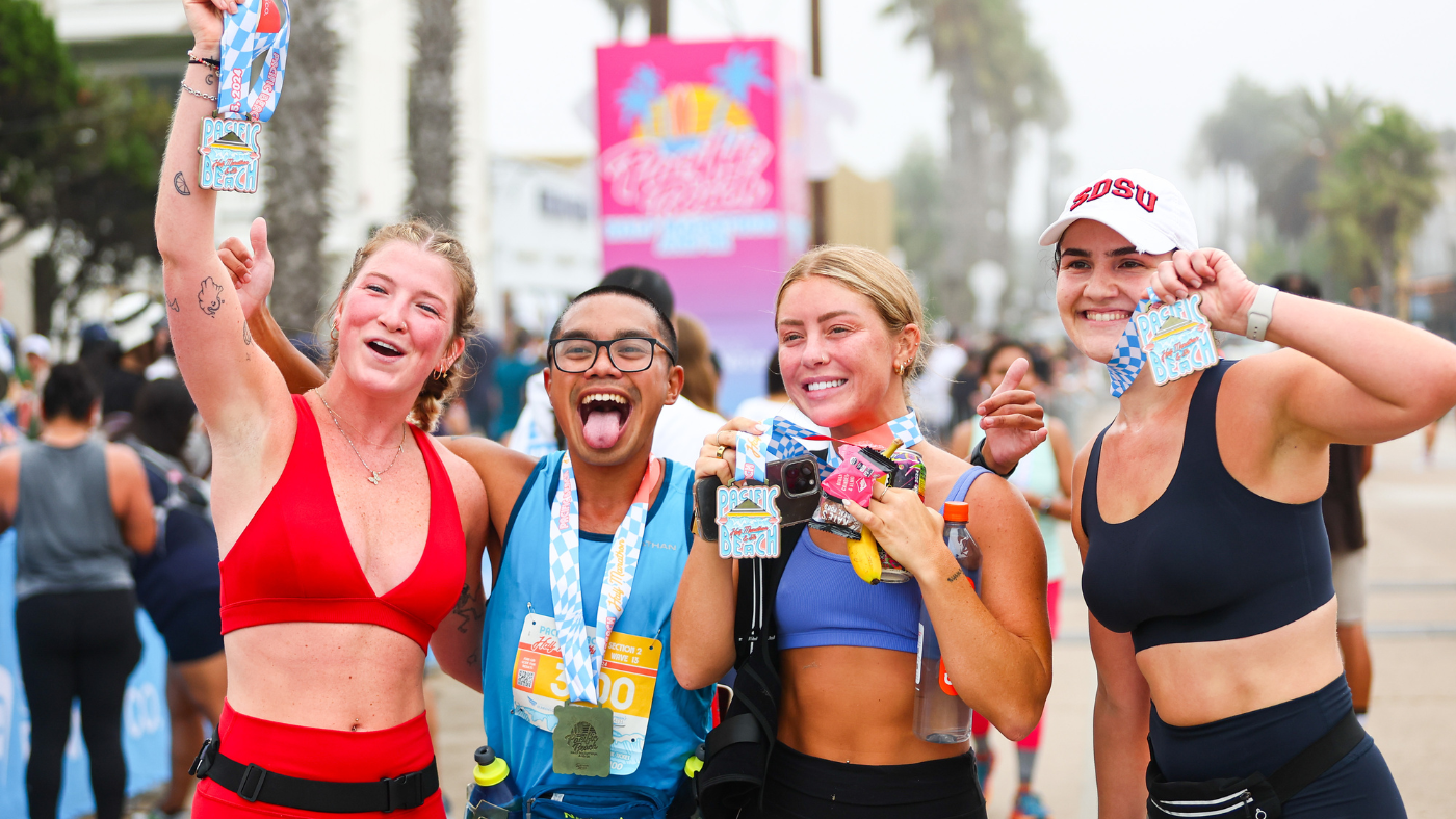 Four smiling runners pose together, holding up race medals at a finish line. They wear athletic gear and appear happy and energetic, with palm trees and event signs in the background.