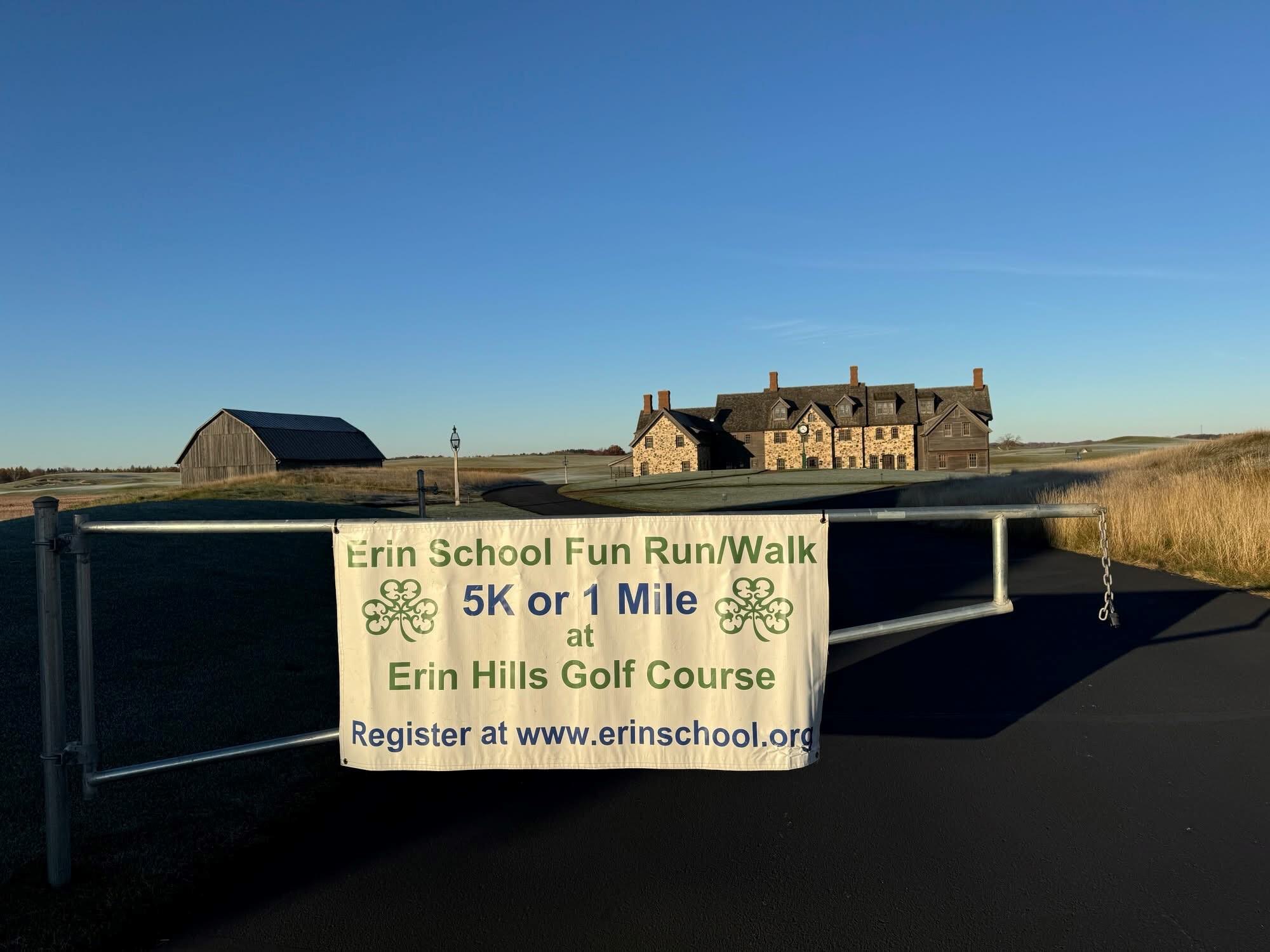 A banner on a gate advertises the Erin School Fun Run/Walk 5K or 1 Mile at Erin Hills Golf Course, with a registration link. A barn and stone building are visible under a clear blue sky.