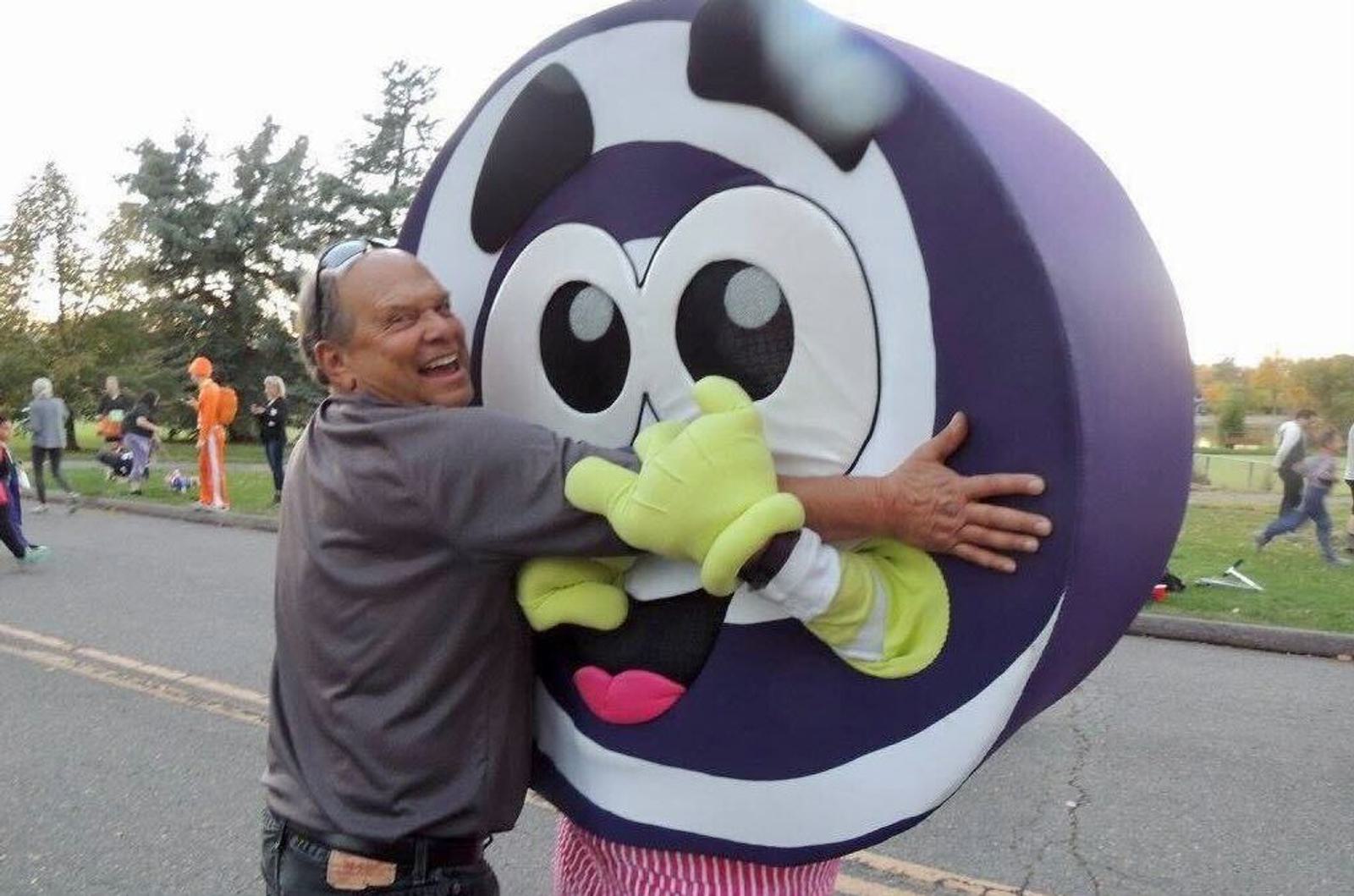 A man smiles and poses as he hugs a person in a large, round, cartoonish costume with big eyes, a wide mouth, and purple and white colors. They are outdoors on a road with trees and people in the background.