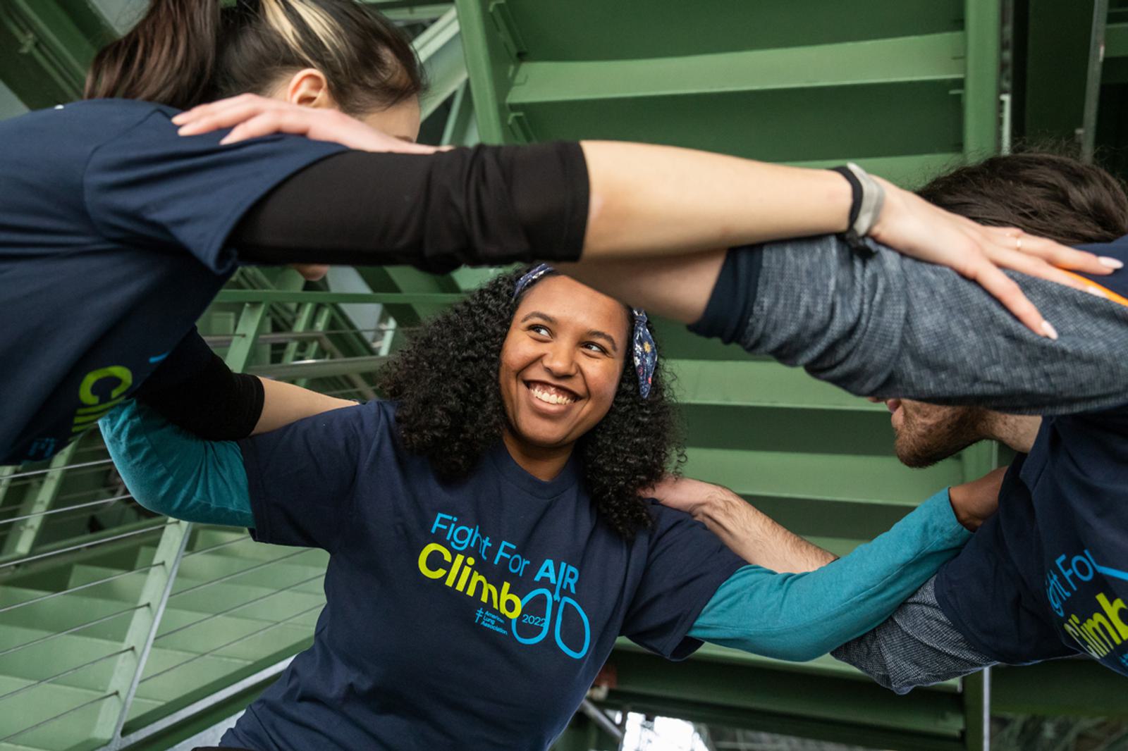 Three people wearing navy "Fight For Air Climb" shirts are forming a circle with their arms linked. The woman in the middle is smiling, under a green metal structure.
