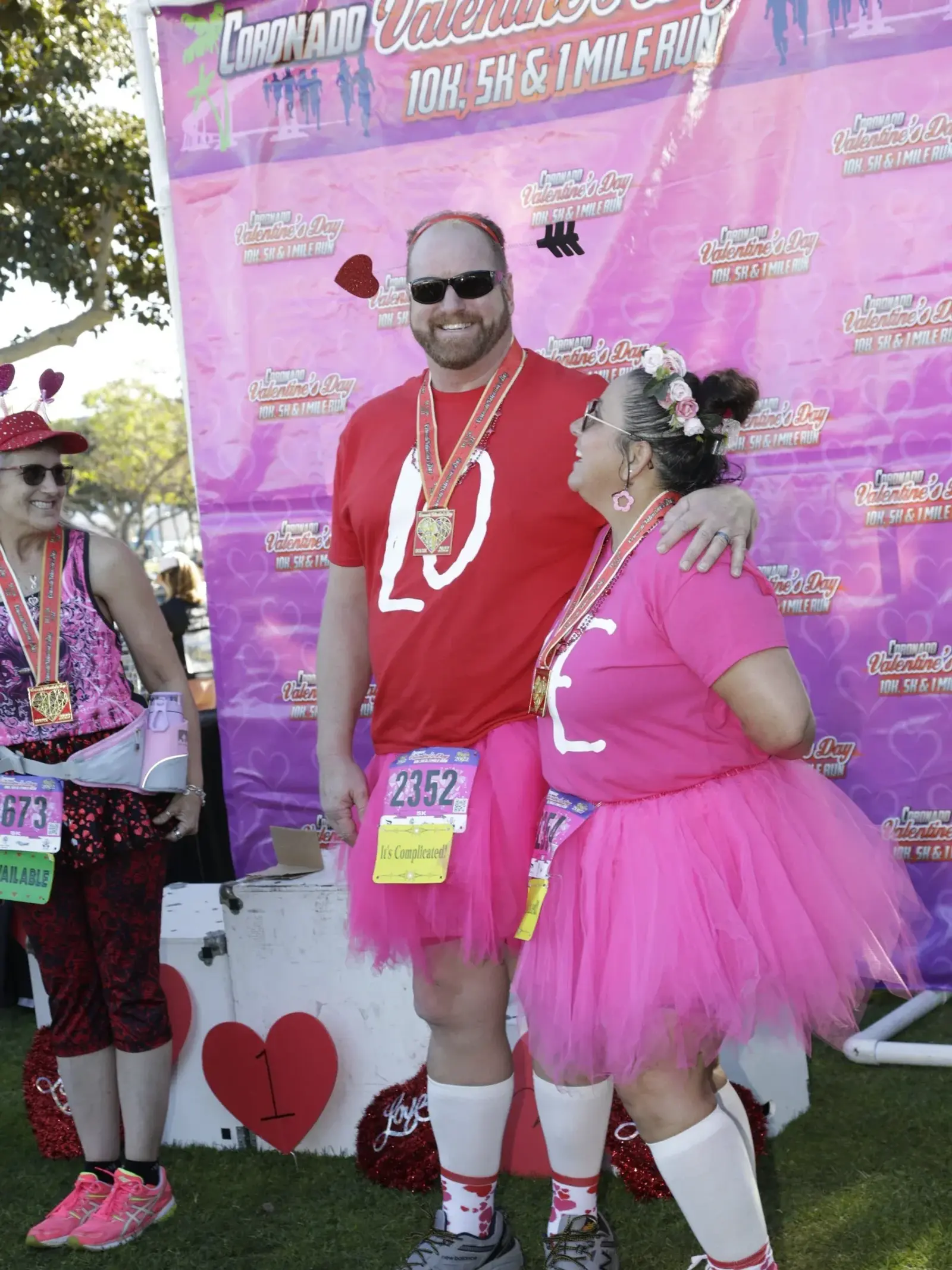 Three people wearing Valentine-themed costumes, medals, and race bibs stand in front of a pink event backdrop at a fun run. Two of them wear matching pink tutus and heart accessories while smiling at each other.
