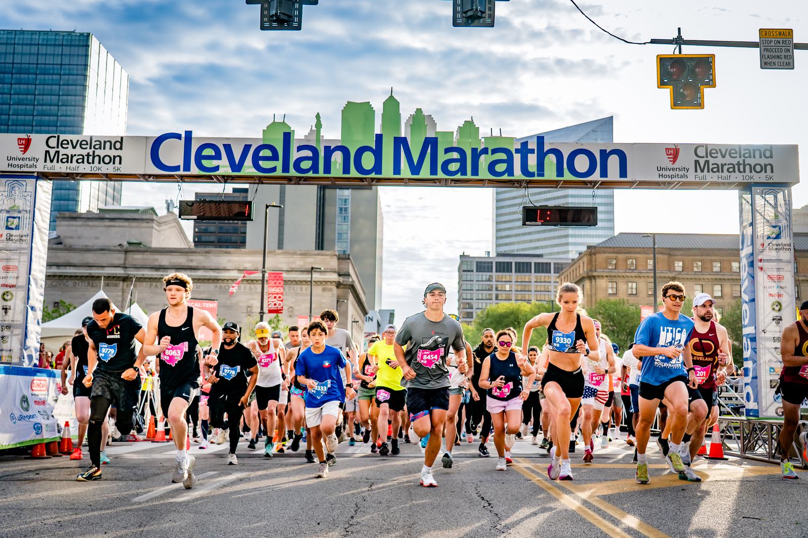 Runners in athletic gear begin the Cleveland Marathon, crossing the start line under a banner with city buildings in the background on a sunny day. Spectators and organizers stand nearby.