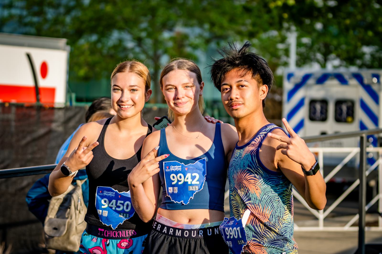 Three young runners stand together smiling and posing with peace signs, wearing race bibs and athletic gear, with trees and an emergency vehicle visible in the background.