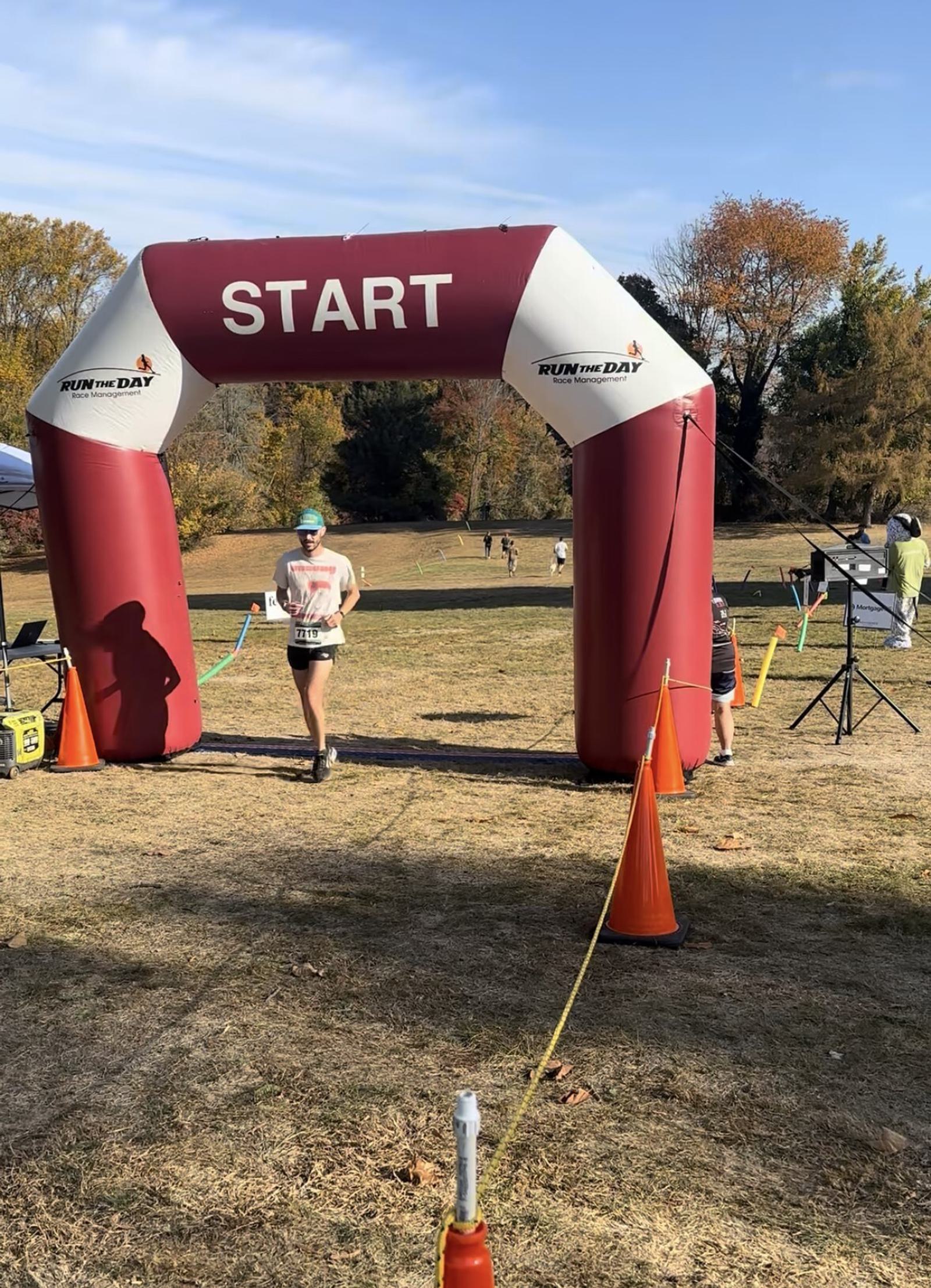 A person in athletic wear runs under a large red and white inflatable arch labeled "START" at an outdoor race event on a sunny day, with trees and other participants visible in the background.