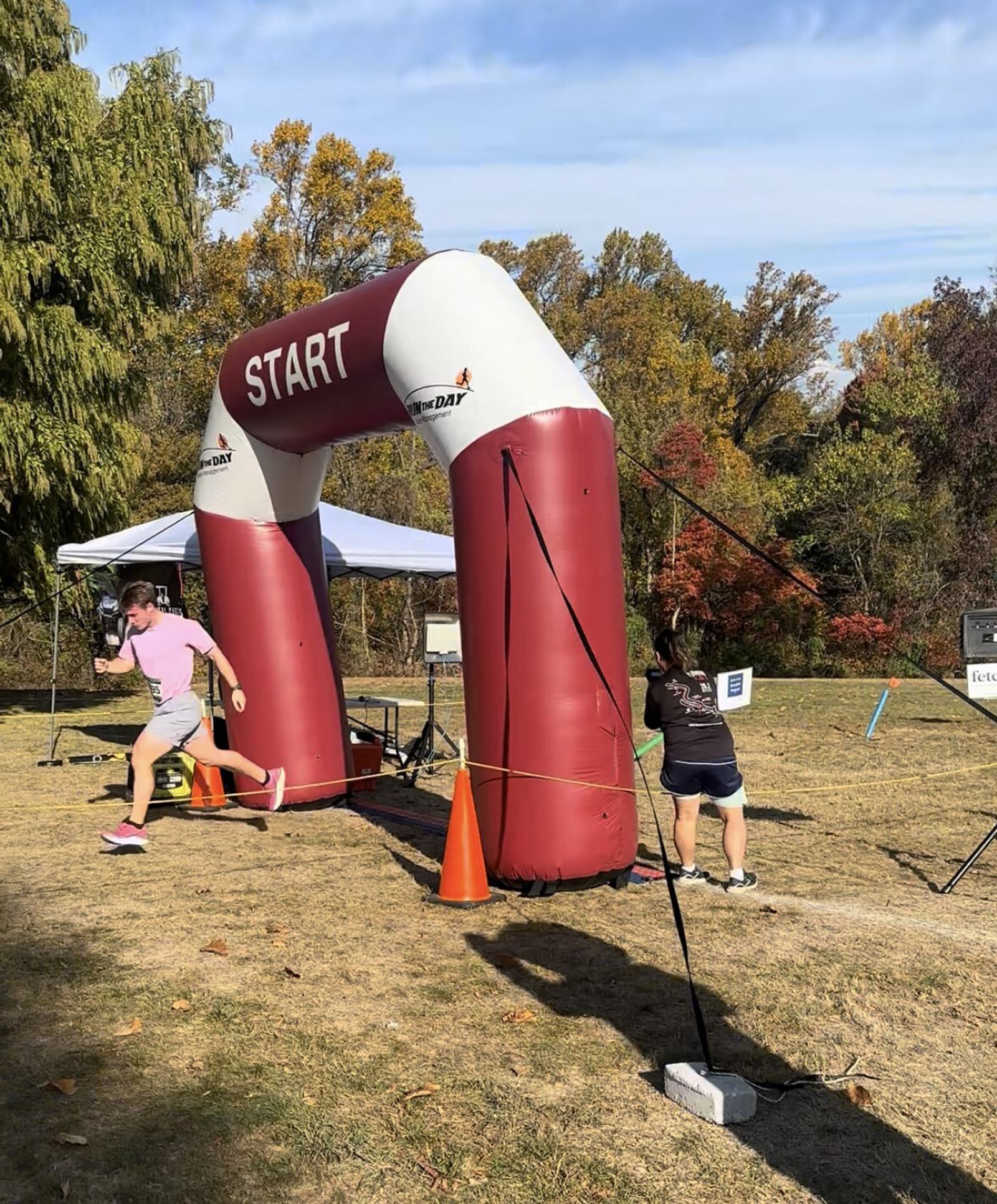 Two people are running under a large inflatable arch labeled "START" at an outdoor event on a sunny day, with trees and grass in the background.