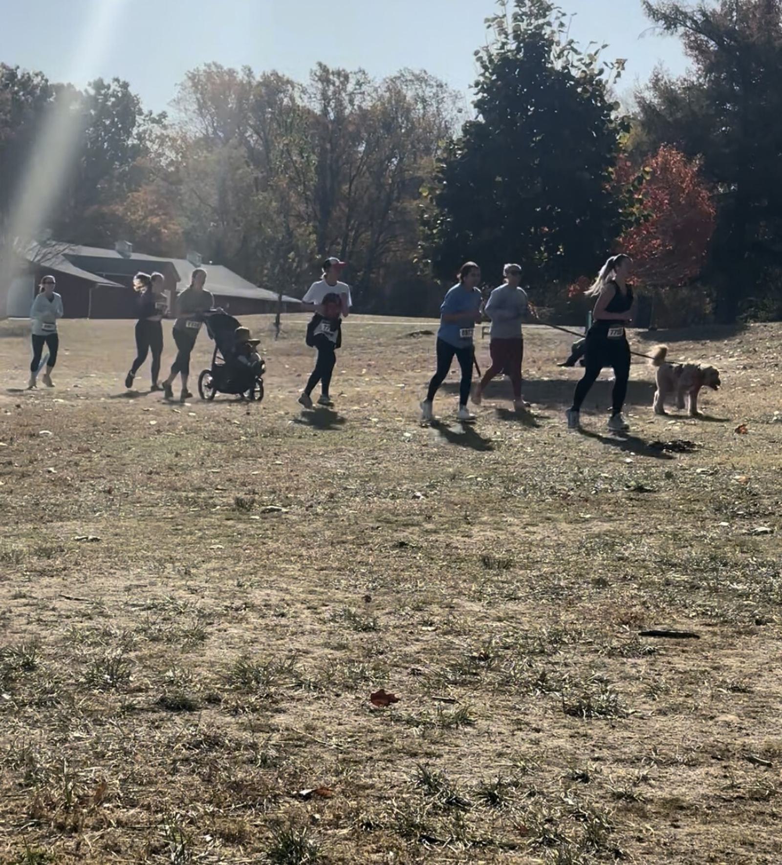 A group of people jogs together across a sunlit park, including a person with a stroller, a child, and someone walking a dog. Trees with autumn foliage are in the background.