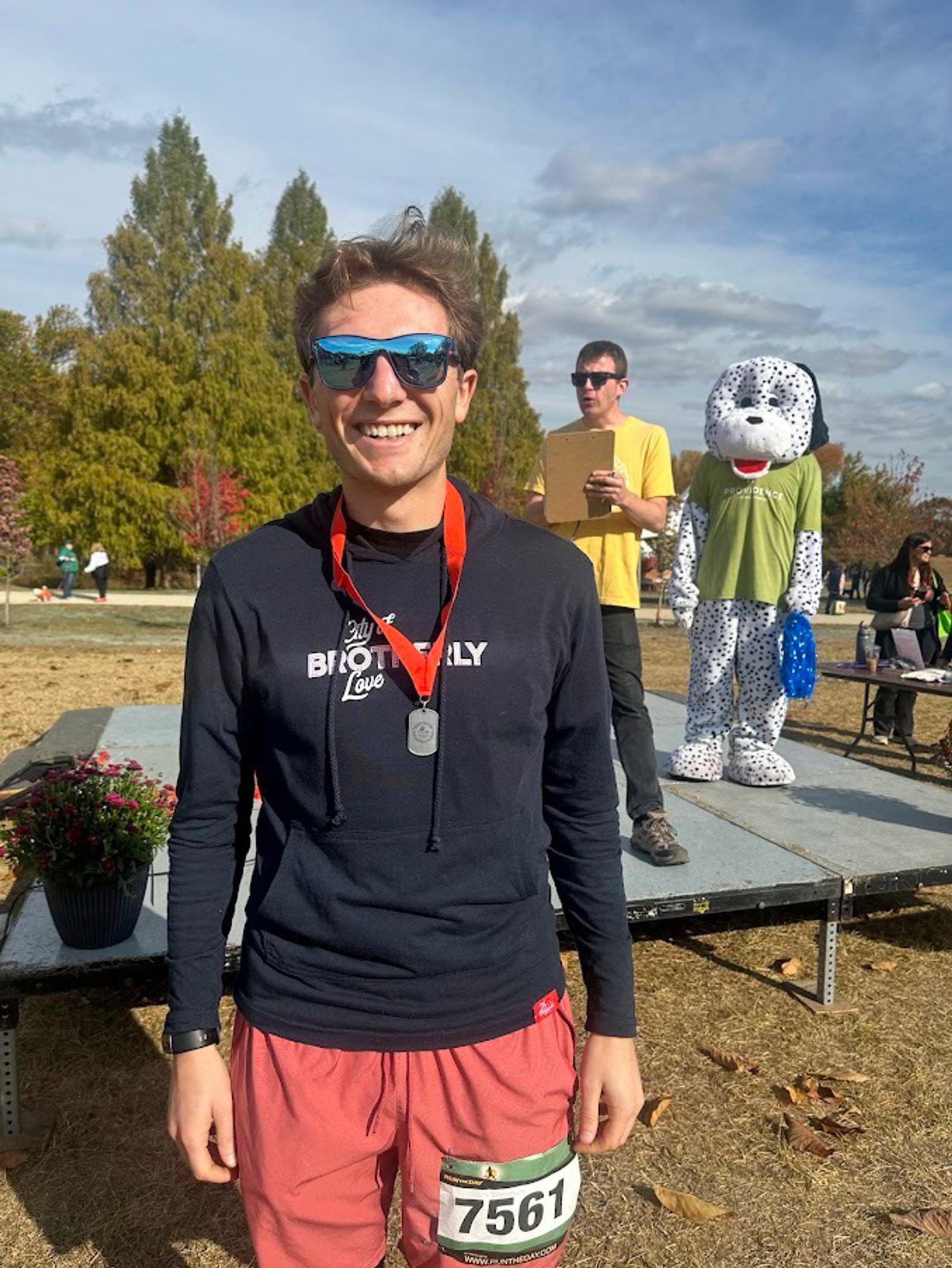 A smiling person wearing sunglasses, a medal, and a "Biotically Love" hoodie stands outdoors at an event. Behind them, a person with a clipboard and someone in a dalmatian mascot costume are on a stage.