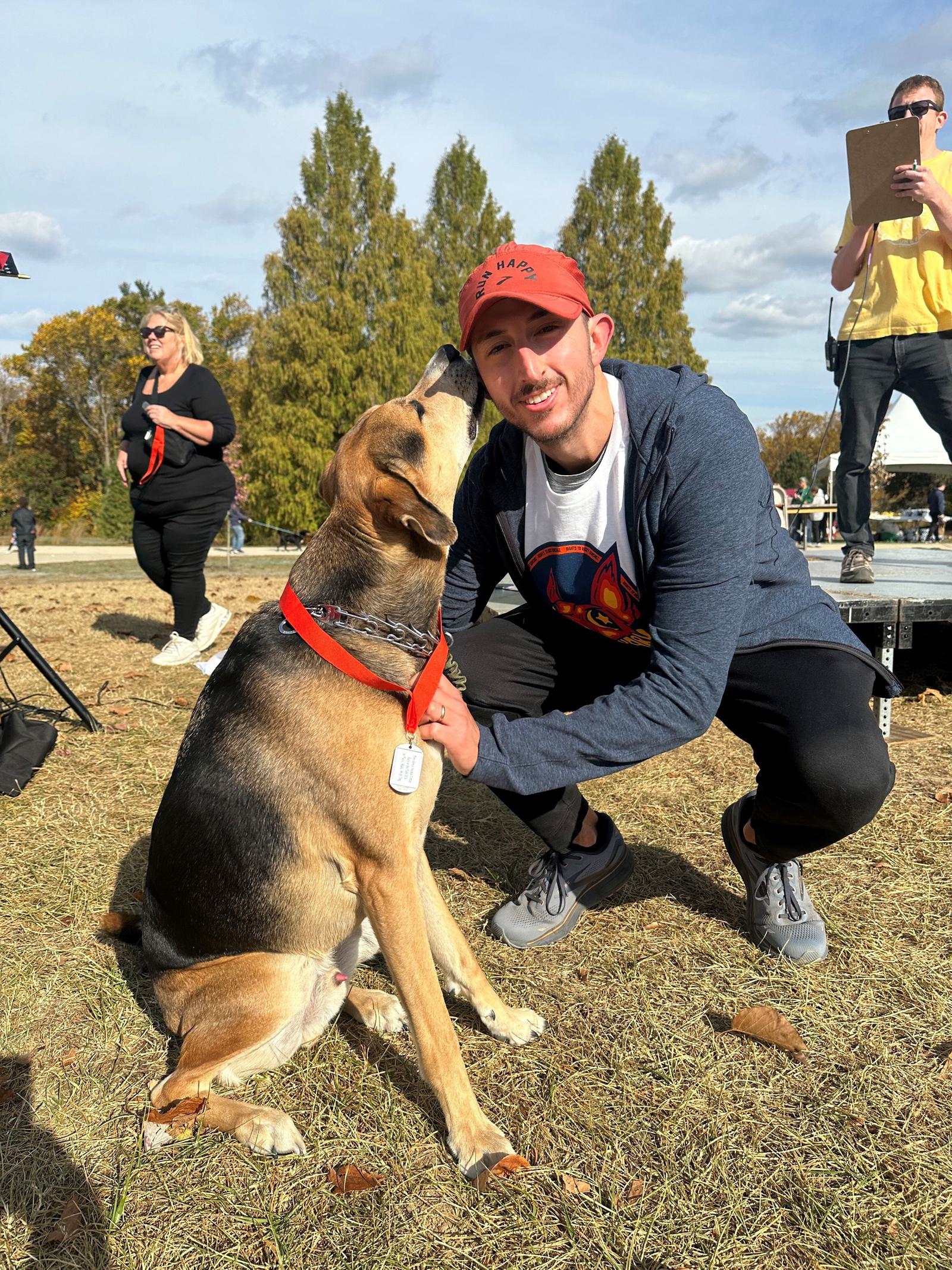 A man in a red cap and blue jacket kneels on grass, smiling as a brown and black dog with a red collar licks his face. People and trees are visible in the background under a partly cloudy sky.