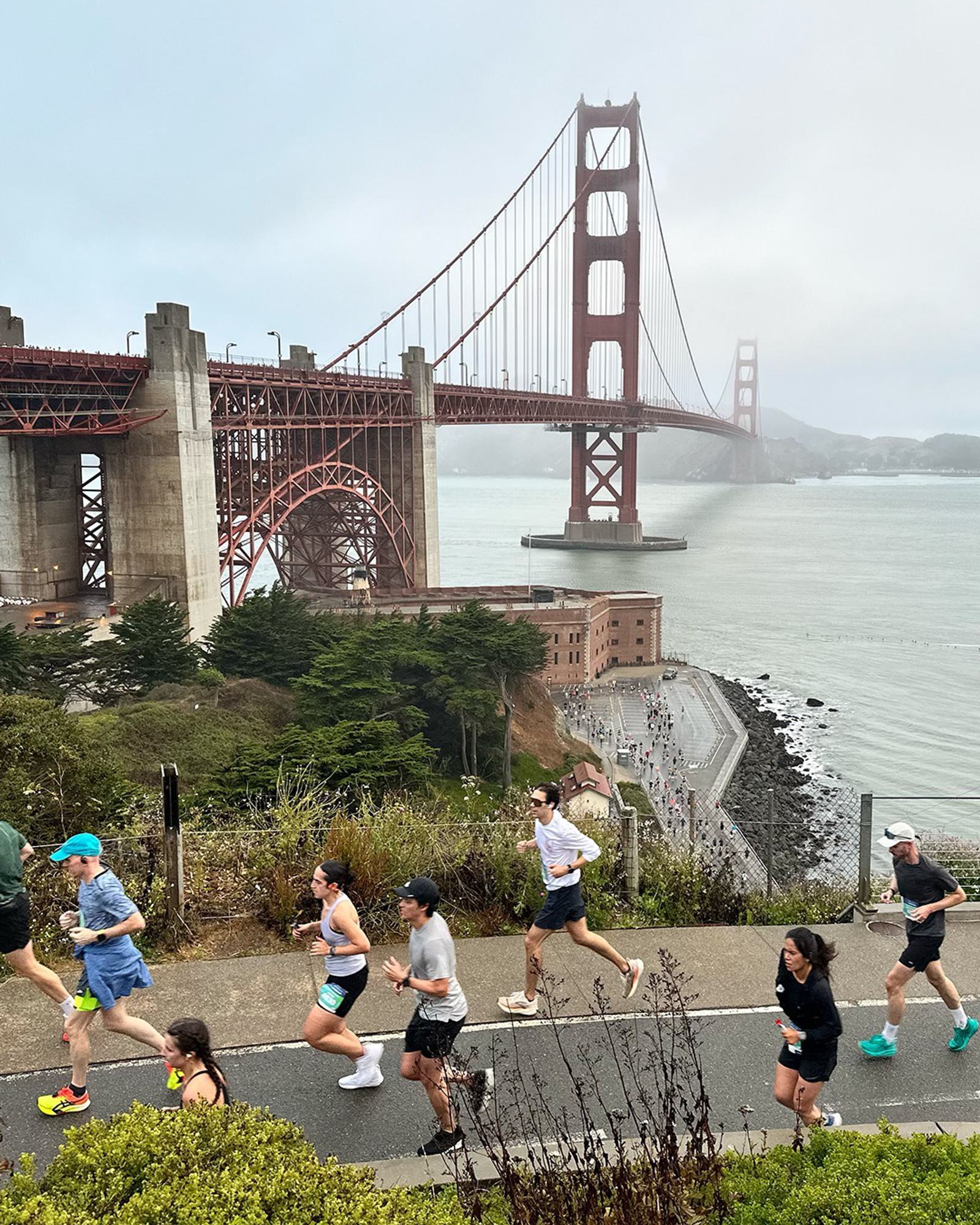 Runners participating in a race jog along a path with the Golden Gate Bridge and San Francisco Bay in the background on a cloudy day.