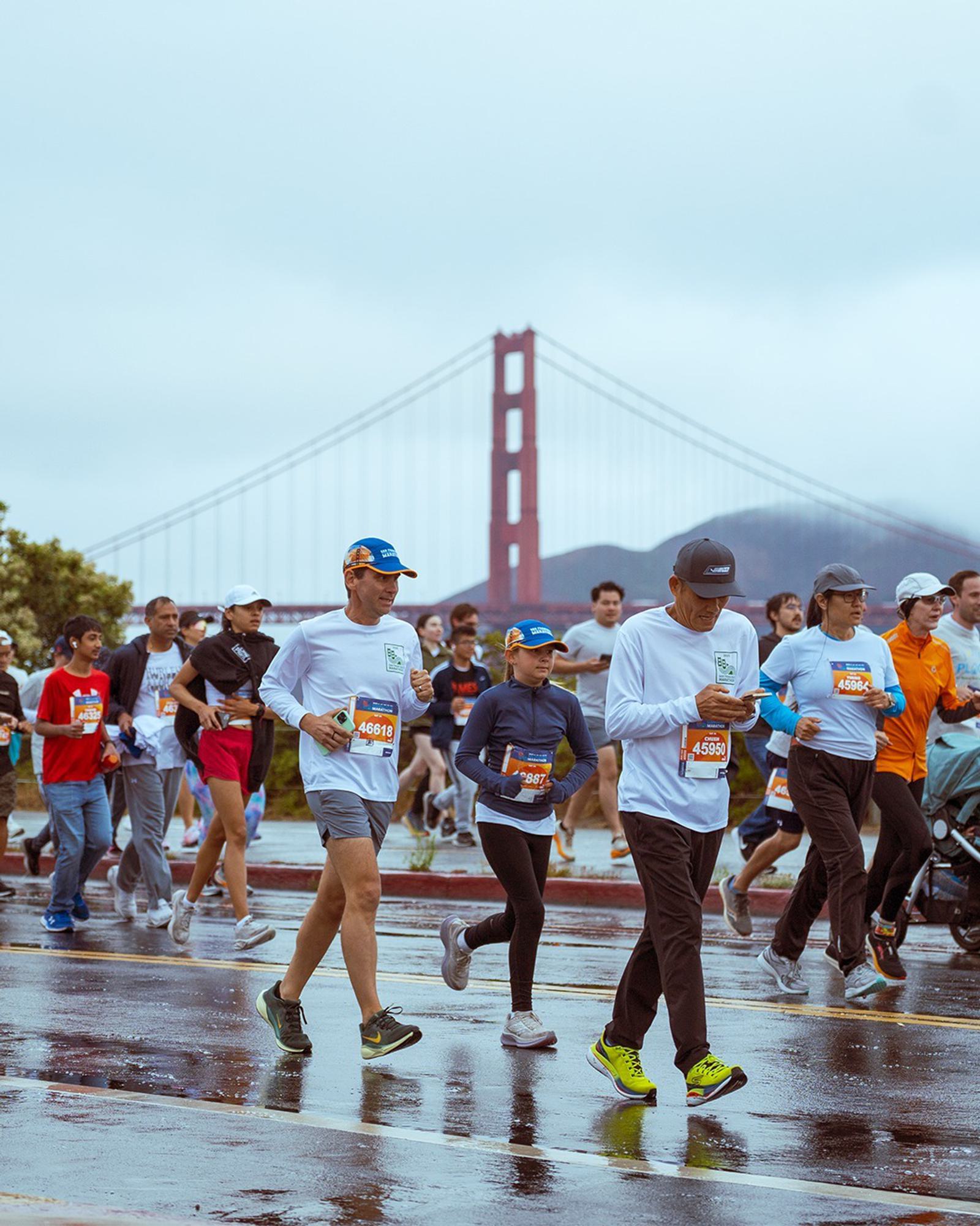 A group of runners, some walking, participate in a race on a wet road with the Golden Gate Bridge visible in the background on a cloudy day.