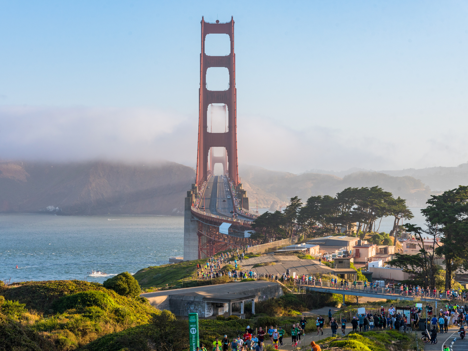 A crowd gathers near the Golden Gate Bridge in San Francisco on a sunny day, with some fog around the hills and the bay in the background. The iconic red bridge stands tall over the water.