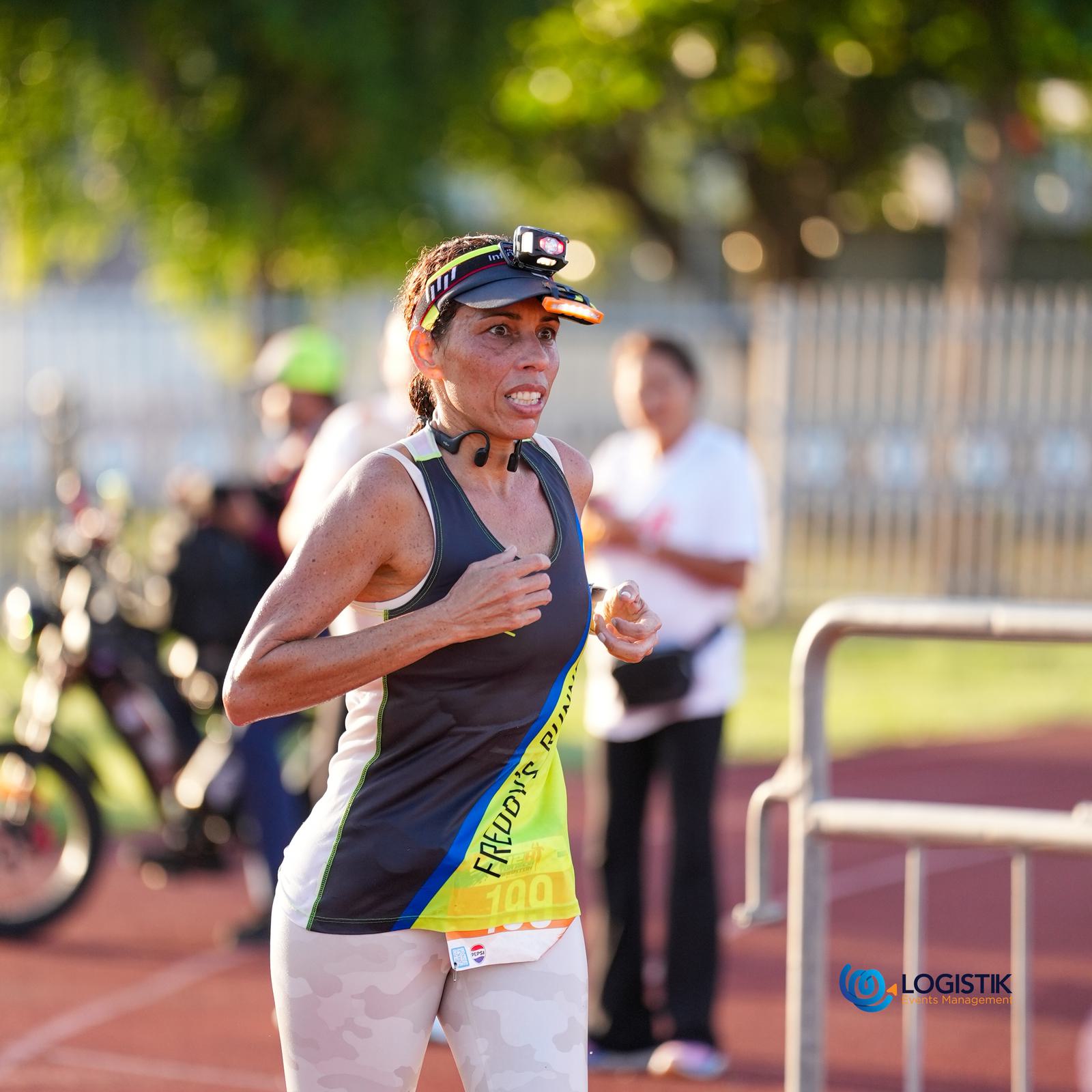 A woman wearing athletic gear and a headlamp runs on a track during a daytime race event, with other participants and a motorcycle visible in the background.