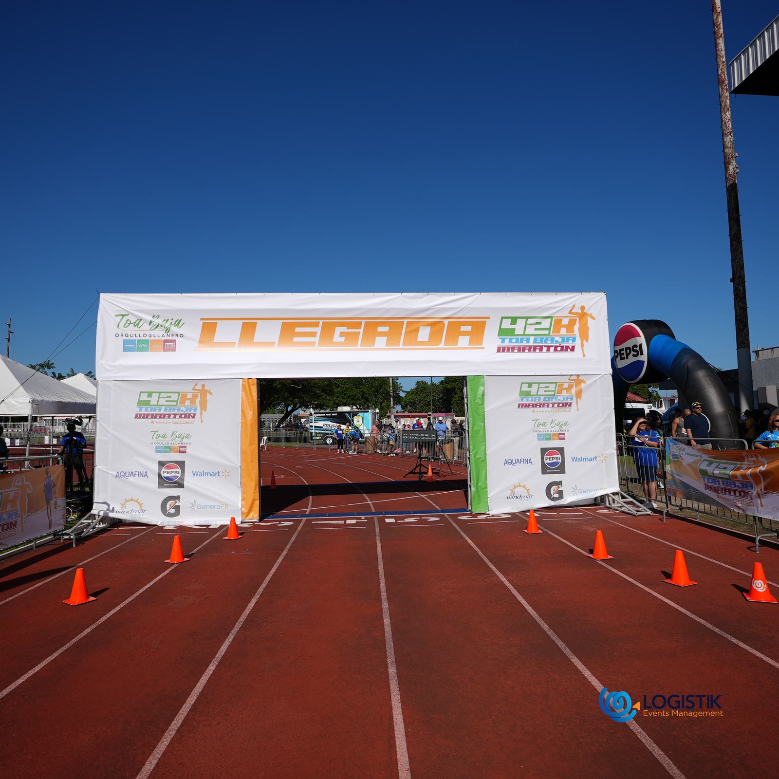 A finish line banner reading "LLEGADA 42K MARATÓN" is set up on a red running track, surrounded by orange cones, with event tents, people, and sponsor logos visible in the background.