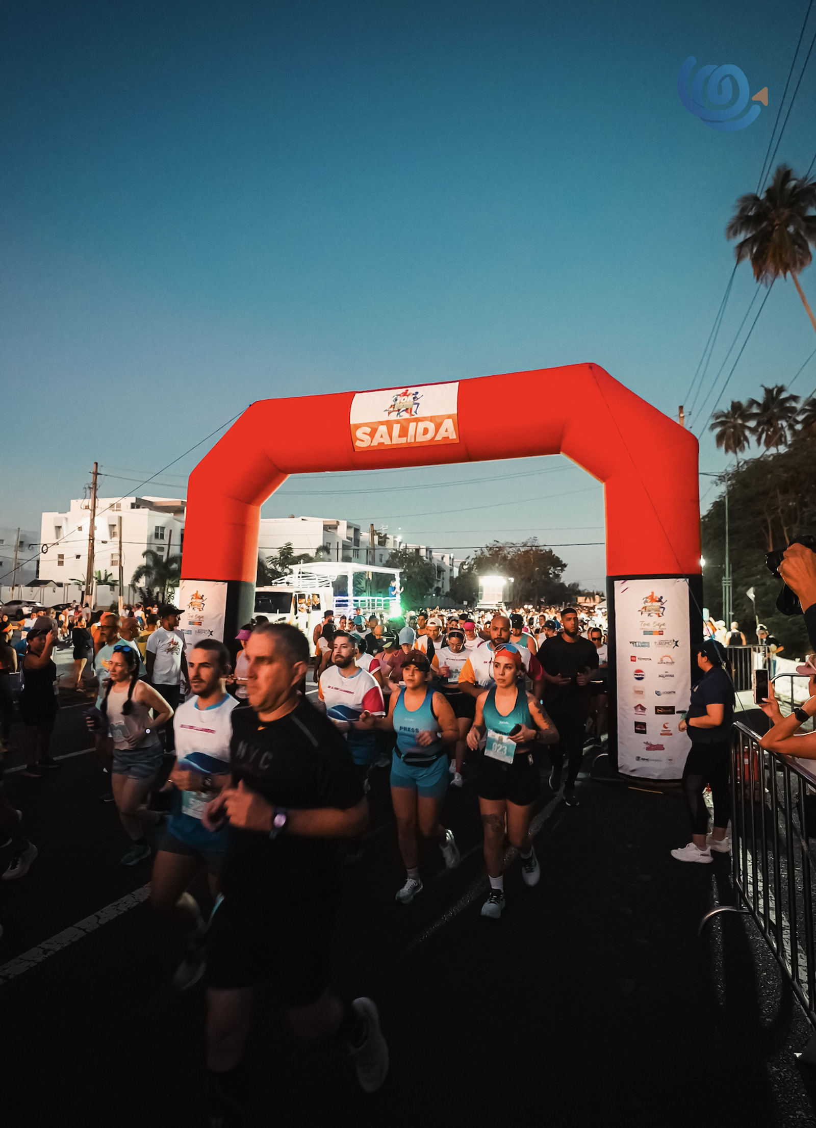 A large group of runners pass under a red inflatable arch labeled "SALIDA" at the start of a race, with buildings, palm trees, and early morning light in the background.
