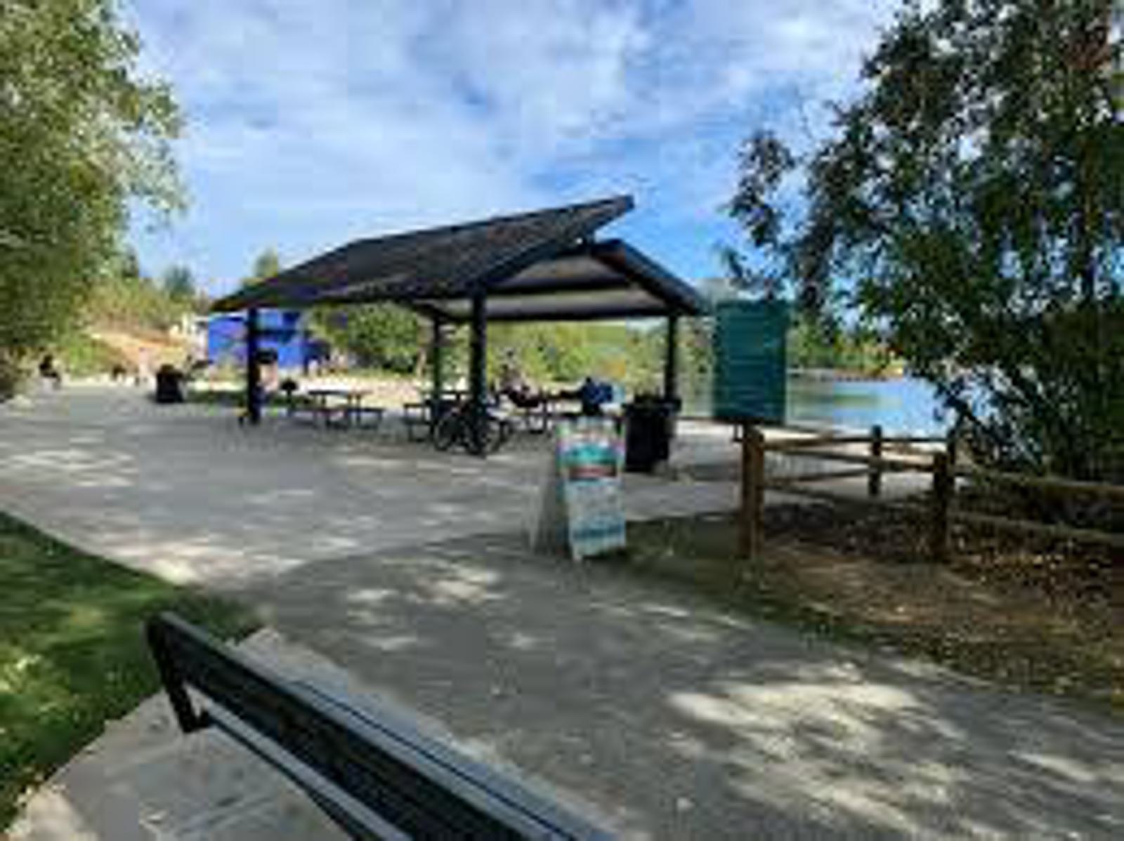 A paved path leads to a covered picnic area with tables near a lake, surrounded by trees. A bench sits in the foreground and a wooden fence lines the walkway under a partly cloudy sky.