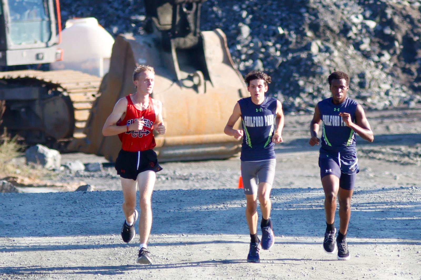 Three young men run on a gravel path at a construction site. One wears a red shirt, the other two wear matching navy and green uniforms. A large excavator and rocks are visible in the background.