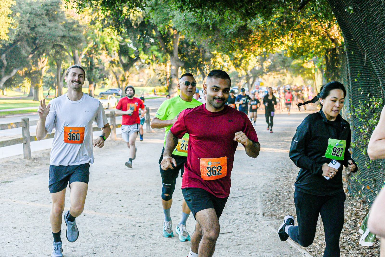 Runners participate in a race on a tree-lined path. One man in front wears a red shirt and number 362, while nearby runners wear race bibs. The scene is lively with people in motion and sunlight filtering through trees.