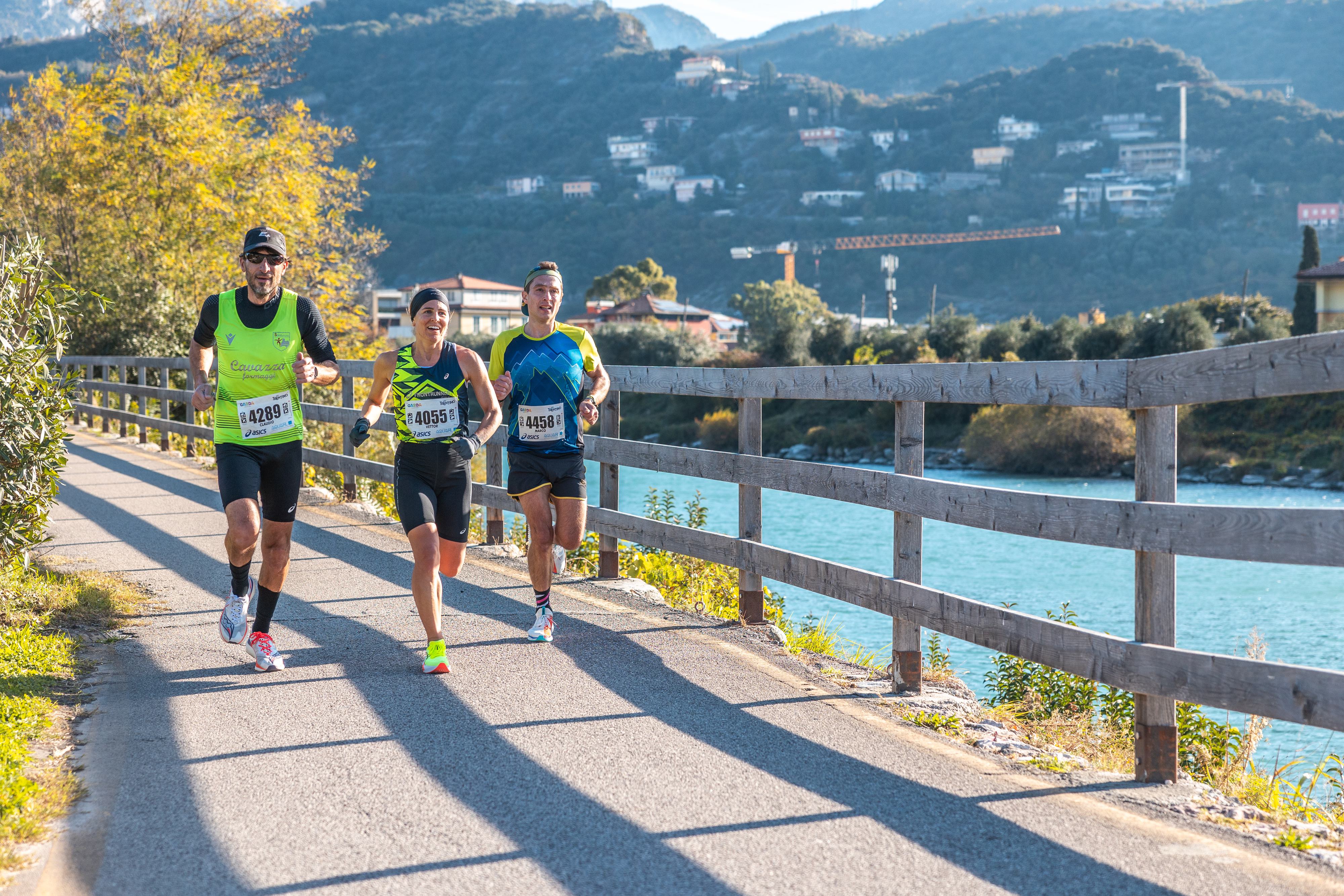 Three runners in athletic gear race along a paved riverside path, with numbered bibs visible. The sun is shining, and mountains, trees, and houses are in the scenic background.