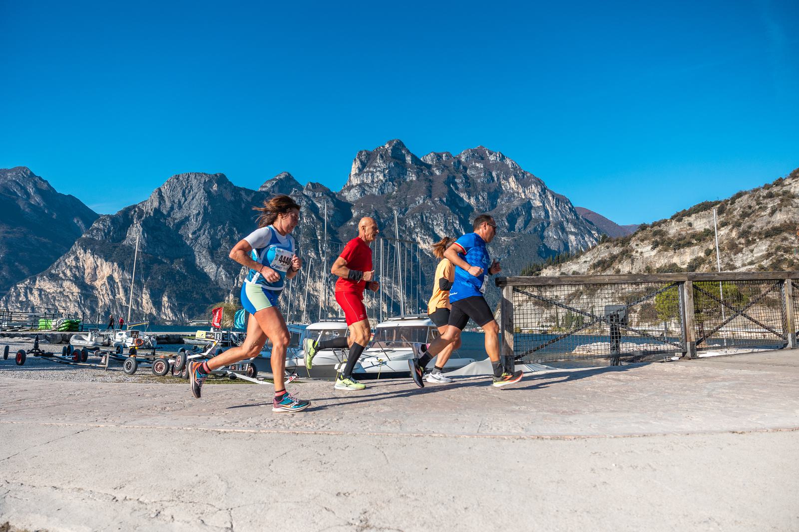 Four people are running together outdoors near a marina, with sailboats and mountains in the background under a clear, blue sky.
