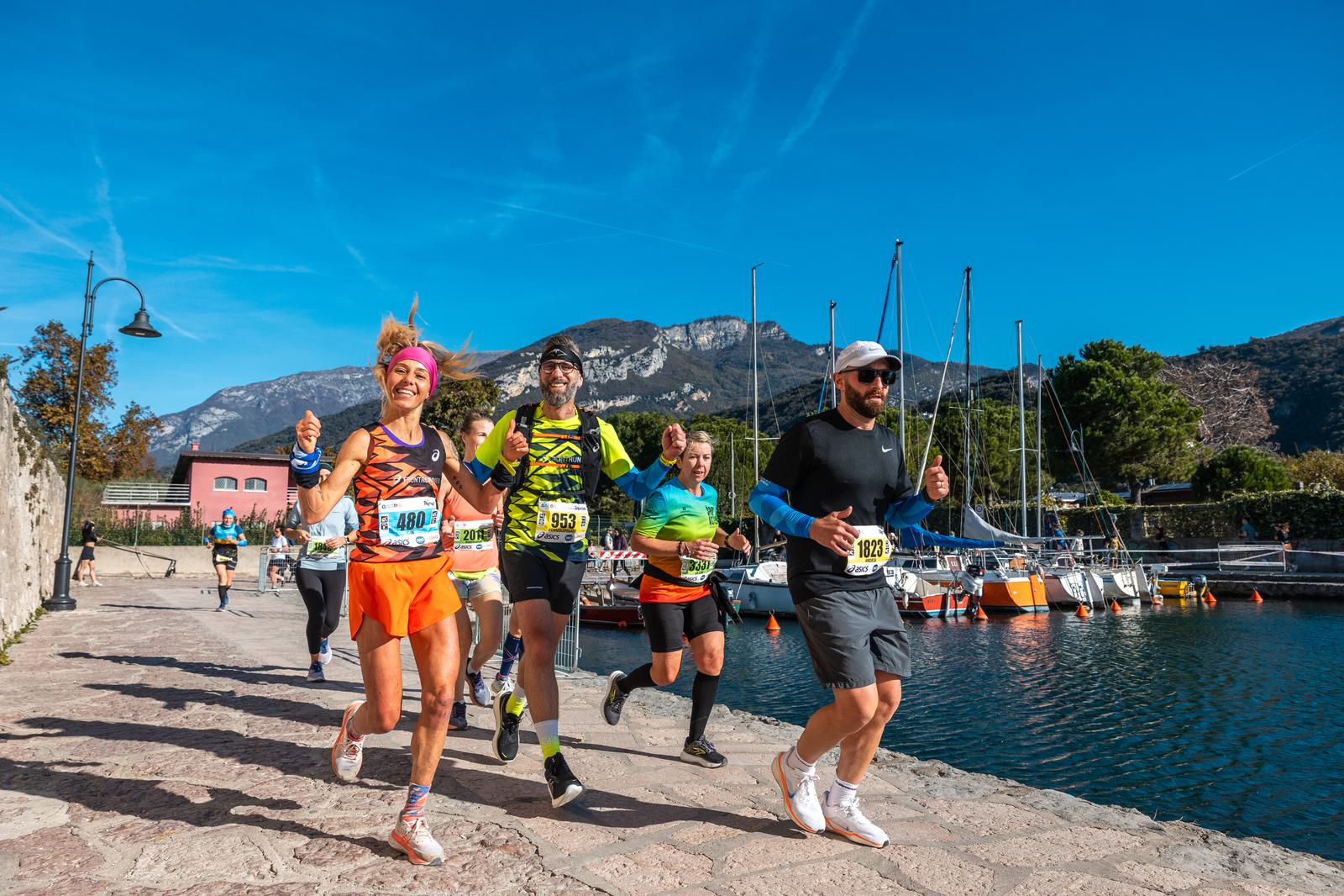 A group of marathon runners, smiling and giving thumbs up, run along a waterfront path with sailboats and mountains in the background under a clear blue sky.