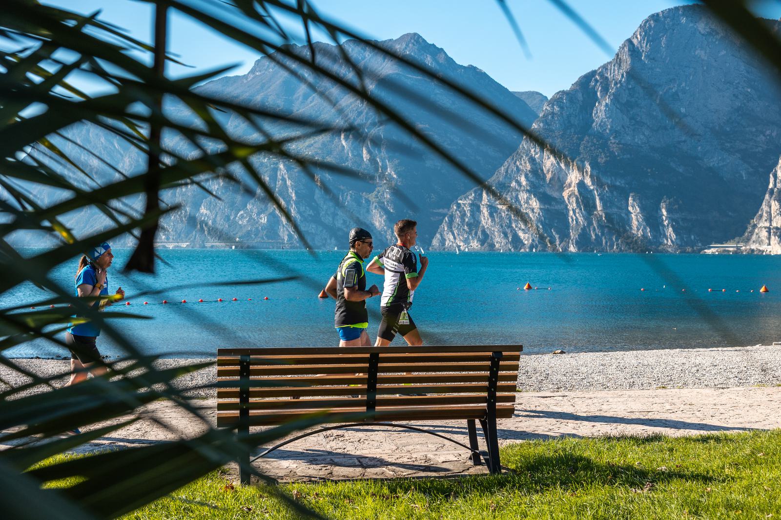 Three runners jog along a lakeside path, passing an empty bench. Blue water and mountains are visible in the background. The scene is framed by out-of-focus palm leaves in the foreground.