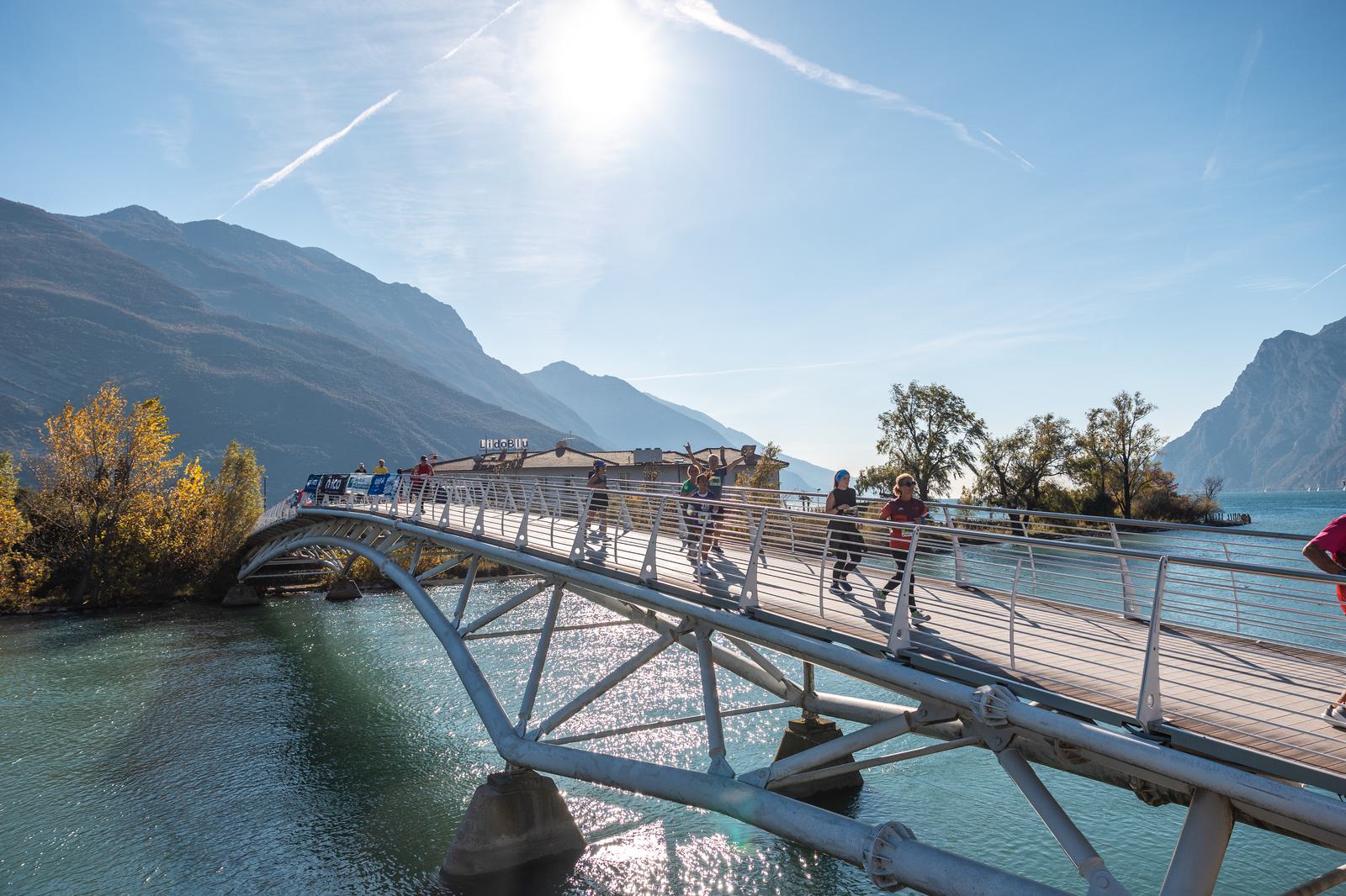 People walk and ride bikes across a modern arched bridge over blue water, with mountains and trees in the background and the sun shining brightly in a clear sky.