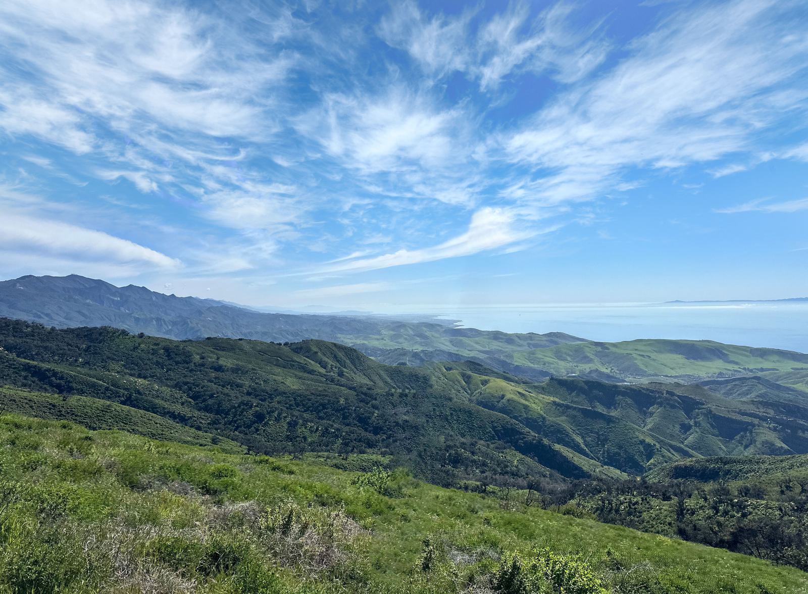 Rolling green hills and mountains stretch toward the distant coastline under a bright blue sky with wispy clouds. The scene is lush, vibrant, and expansive, showcasing natural beauty and open space.