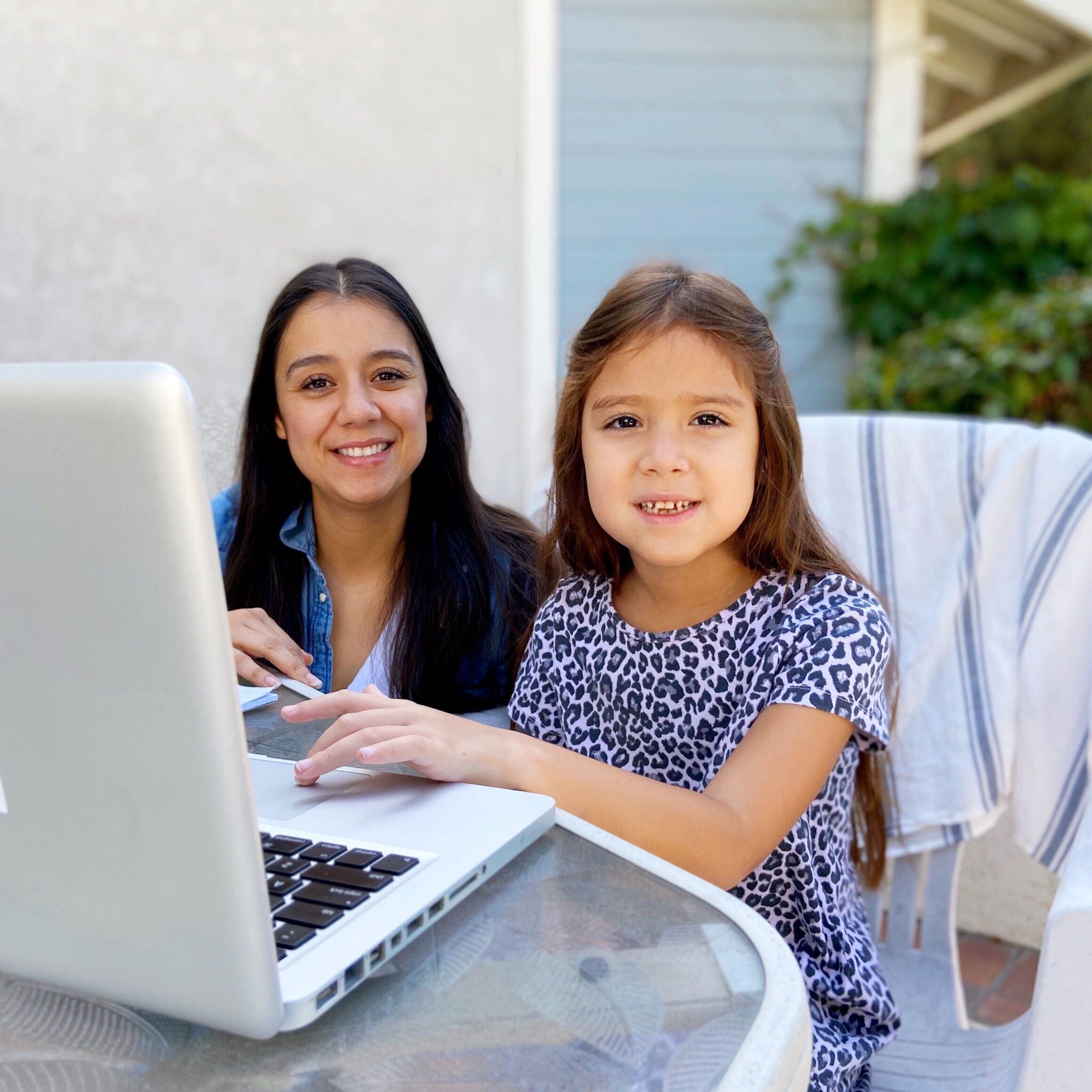 A young woman and a girl sit at a glass table outdoors, smiling at the camera. The girl is in front of a laptop, with greenery and a light-colored house in the background.