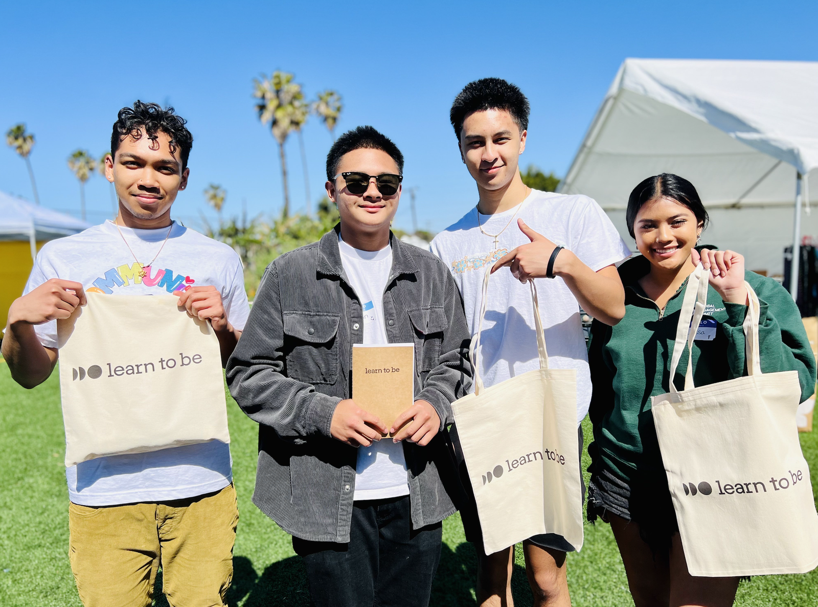 Four young adults stand outside on grass, smiling and holding "Learn To Be" tote bags and a notebook, with tents and palm trees visible in the sunny background.