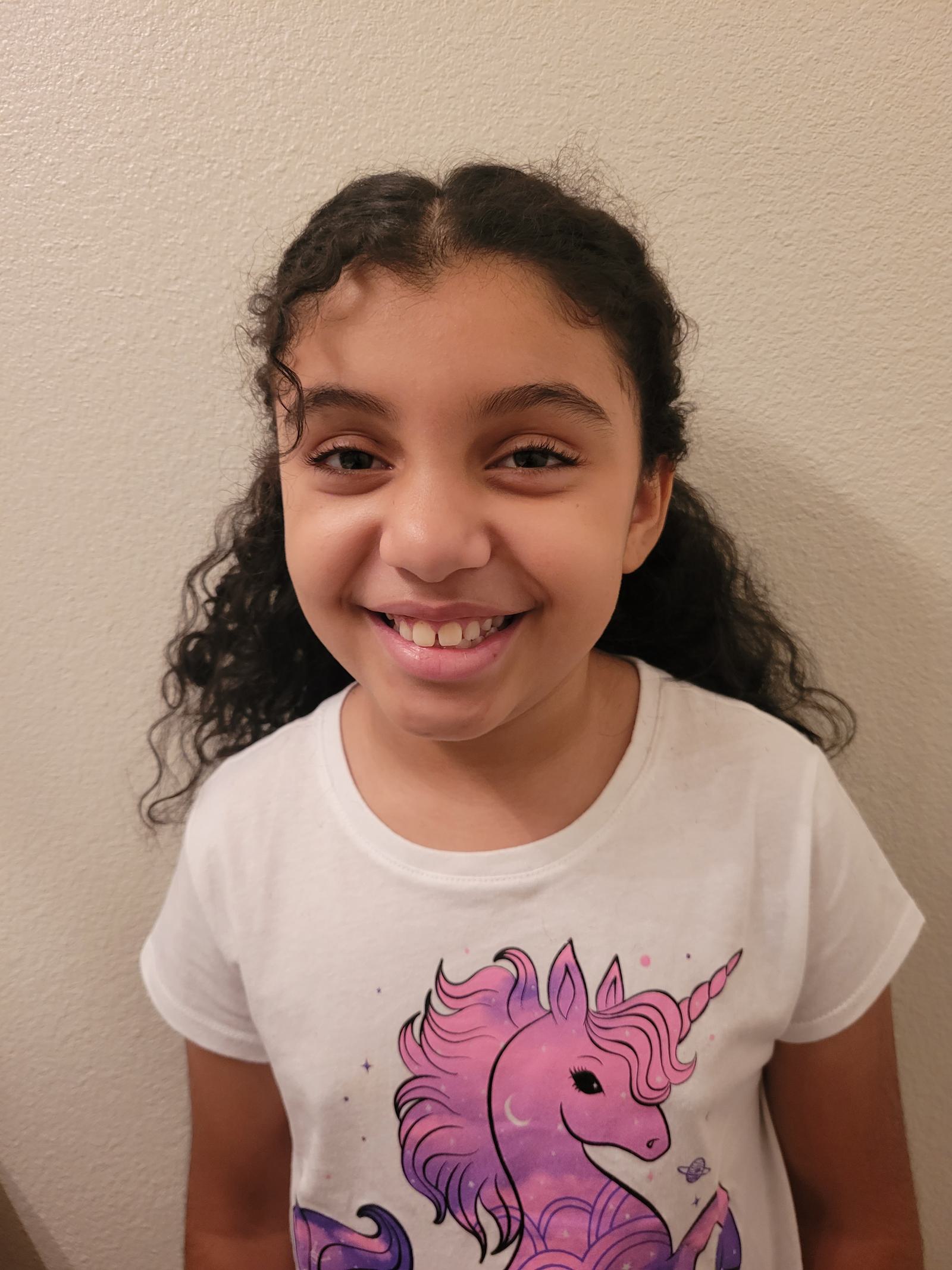 A smiling young girl with curly hair stands against a light-colored wall, wearing a white shirt featuring a colorful unicorn graphic.